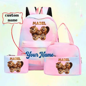 May include: A three-piece set of school bags in pink and blue tie-dye. The set includes a backpack, lunch bag, and pencil case, each featuring a cartoon beaver family and the name "MABEL". The backpack and pencil case have the text "Your Name".