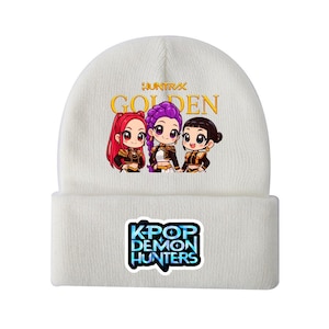 Könnte beinhalten: Weißer Beanie mit einer bunten Grafik von drei Cartoon-Charakteren und dem Text "HUNTRAX GOLDEN". Darunter steht "KPOP DEMON HUNTERS" in Blau und Weiß. Ein Winteraccessoire für K-Pop-Fans.