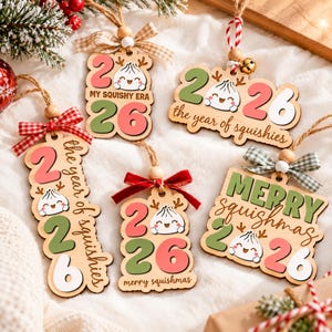 Può includere: Cinque ornamenti in legno con l'anno 2026, un personaggio a forma di gnocco dei cartoni animati e testi tra cui "MY SQUISHY ERA", "the year of squishies" e "merry squishmas". Ogni ornamento è decorato con una perlina di legno e un fiocco.