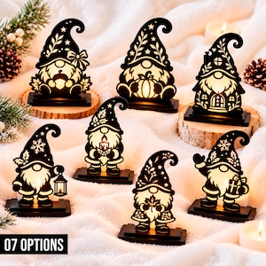Op de afbeelding: Zeven zwarte kabouterfiguren in silhouet met warme interne verlichting, elk gemonteerd op een rechthoekige voet. De ontwerpen bevatten diverse seizoensgebonden motieven zoals pompoenen, lantaarns, cadeaus en bladerpatronen.