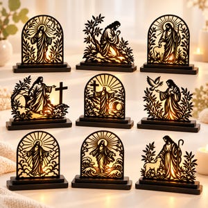 Könnte beinhalten: Eine Sammlung von neun dekorativen Leuchtdisplays in schwarzer Silhouette mit verschiedenen religiösen Szenen von Jesus, die jeweils auf einem rechteckigen Sockel montiert sind und durch eine interne warme Lichtquelle die filigranen Ausschnitte beleuchten.