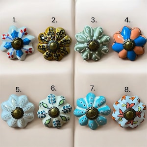 Könnte beinhalten: Acht dekorative Keramik-Schrankknöpfe in Blumenform. Jeder Knopf hat ein anderes Farbschema, darunter Blau, Orange, Grün und Gold, mit einer messingfarbenen Mitte. Die Knöpfe sind in zwei Reihen zu je vier Stück angeordnet.