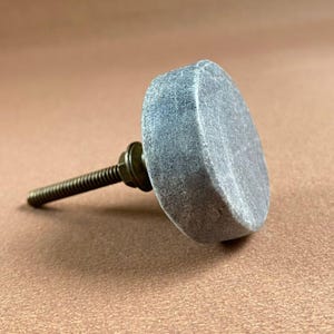 Pode incluir: Um puxador de arm&aacute;rio de pedra cinza com um parafuso e arruela de lat&atilde;o. O puxador tem formato quadrado arredondado. A ferragem &eacute; para m&oacute;veis como gavetas e arm&aacute;rios.