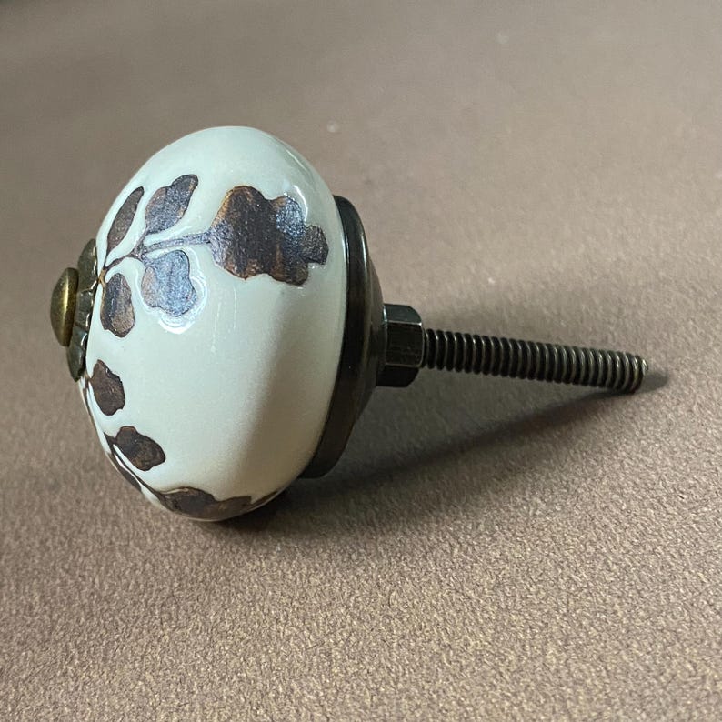 Puede incluir: Pomo de armario de cer&aacute;mica color crema con un dise&ntilde;o floral marr&oacute;n oscuro. El pomo tiene una base de metal y un perno roscado para la instalaci&oacute;n. El herraje tiene una est&eacute;tica vintage.