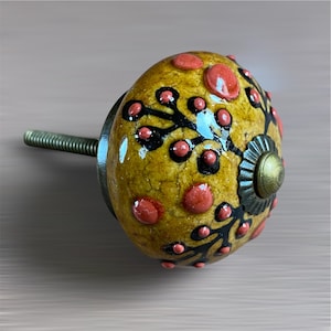 May include: A decorative cabinet knob with a yellow base, decorated with red and black floral patterns. The knob has a brass-coloured metal base and a threaded rod for installation. The design features raised red dots and black, stylised floral elements.
