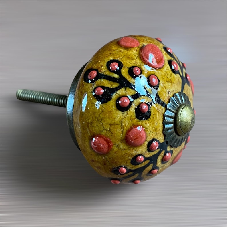 May include: A decorative cabinet knob with a yellow base, decorated with red and black floral patterns. The knob has a brass-coloured metal base and a threaded rod for installation. The design features raised red dots and black, stylised floral elements.