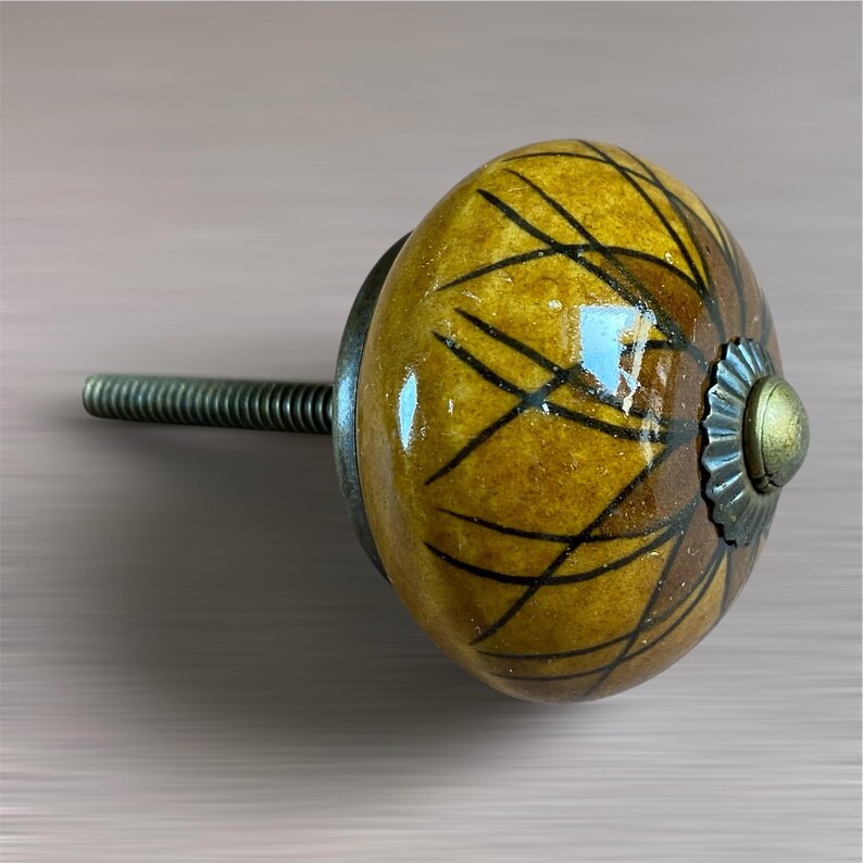 May include: A decorative cabinet knob with a yellow and brown patterned design. The knob has a metal base and a threaded rod for installation. The design features black lines over a yellow and brown background.