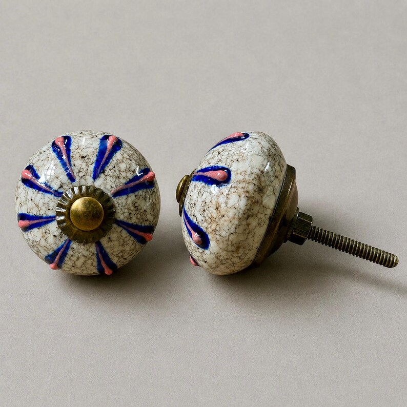 Op de afbeelding: Twee decoratieve keramische kastknoppen. De knoppen zijn wit met een gespikkeld patroon en versierd met blauwe en roze bloemmotieven. Elke knop heeft een messingkleurige basis en een metalen schroefdraad voor installatie.