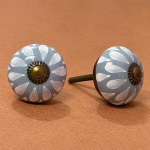 May include: Two round ceramic cabinet knobs featuring a light blue base with a white petal or teardrop pattern radiating from the center. Each knob has a dark brass-toned metal center cap with a scalloped edge and a threaded metal bolt attached.