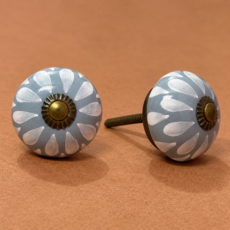 May include: Two round ceramic cabinet knobs featuring a light blue base with a white petal or teardrop pattern radiating from the center. Each knob has a dark brass-toned metal center cap with a scalloped edge and a threaded metal bolt attached.