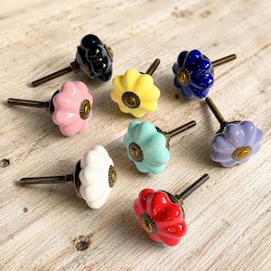 Pode incluir: Uma coleção de oito puxadores de gaveta em cerâmica em várias cores, incluindo preto, rosa, amarelo, azul escuro, azul claro, branco, vermelho e lavanda. Cada puxador tem formato de flor com um parafuso central e ferragens em latão.