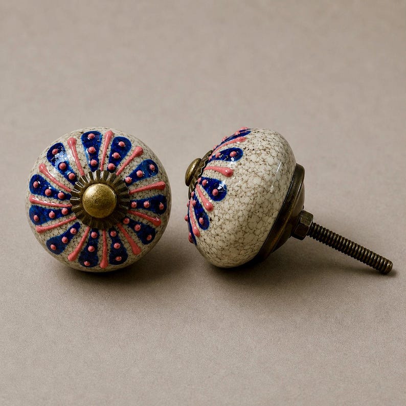 Op de afbeelding: Twee decoratieve keramische kastknoppen. De knoppen zijn gebroken wit met een gespikkeld patroon en hebben een blauw en roze bloemmotief. Elke knop heeft een messingkleurige basis en een schroefdraad voor installatie.