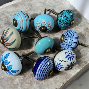 Puede incluir: Una colección de pomos de cerámica decorativos en varios tonos de azul, blanco y turquesa. Cada pomo presenta diseños únicos pintados a mano, incluyendo patrones florales y rayas. Los pomos tienen herrajes de color latón.