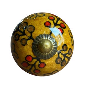 May include: A decorative cabinet knob with a yellow base, decorated with red and brown floral patterns and black outlines. The knob has a brass-coloured metal base and a central screw. This unique hardware adds elegance to any furniture piece.