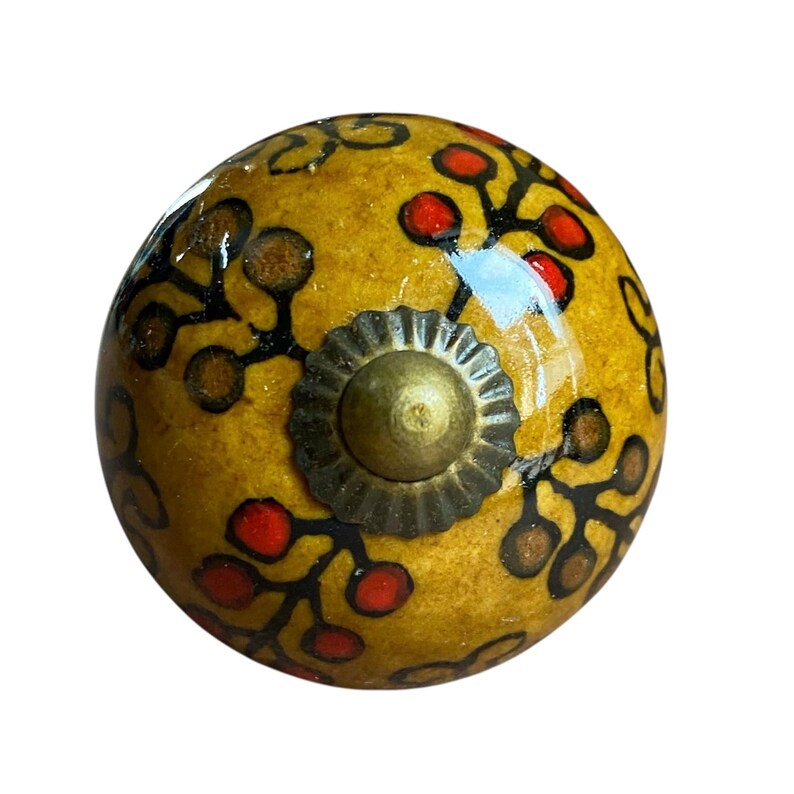 May include: A decorative cabinet knob with a yellow base, decorated with red and brown floral patterns and black outlines. The knob has a brass-coloured metal base and a central screw. This unique hardware adds elegance to any furniture piece.