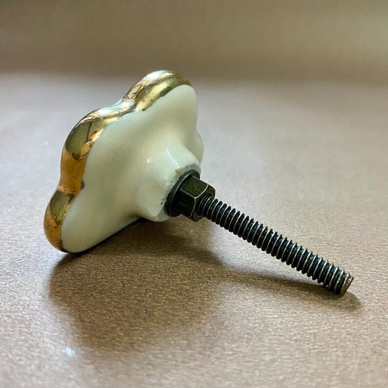 May include: A decorative cabinet knob with a white, floral-shaped ceramic face and gold trim. The knob is attached to a black threaded bolt with a hex nut. The knob is designed for furniture and home decor.