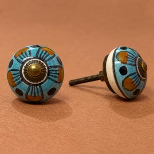 May include: Two round ceramic cabinet knobs featuring a light blue base with a hand-painted floral pattern in dark blue and mustard yellow. Each knob has a central brass-toned metal accent and a threaded metal bolt extending from the back.