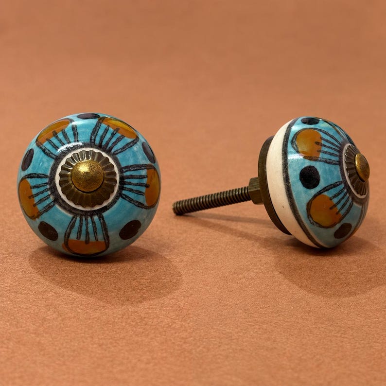 May include: Two round ceramic cabinet knobs featuring a light blue base with a hand-painted floral pattern in dark blue and mustard yellow. Each knob has a central brass-toned metal accent and a threaded metal bolt extending from the back.