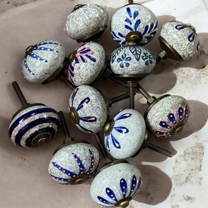 Op de afbeelding: Een verzameling keramische kastknoppen met diverse ontwerpen. De knoppen hebben een gespikkelde, gebroken witte basis met blauwe en roze bloempatronen. Sommige hebben blauwe strepen. Elke knop heeft een messingkleurige basis en schroef.