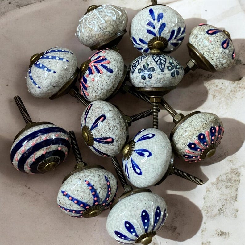 Op de afbeelding: Een verzameling keramische kastknoppen met diverse ontwerpen. De knoppen hebben een gespikkelde, gebroken witte basis met blauwe en roze bloempatronen. Sommige hebben blauwe strepen. Elke knop heeft een messingkleurige basis en schroef.
