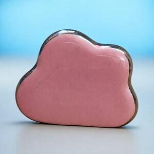 May include: A pink, cloud-shaped gua sha tool with a silver trim. The tool is made of a smooth, glossy material and is designed for facial massage. The background is a gradient of light blue and white.