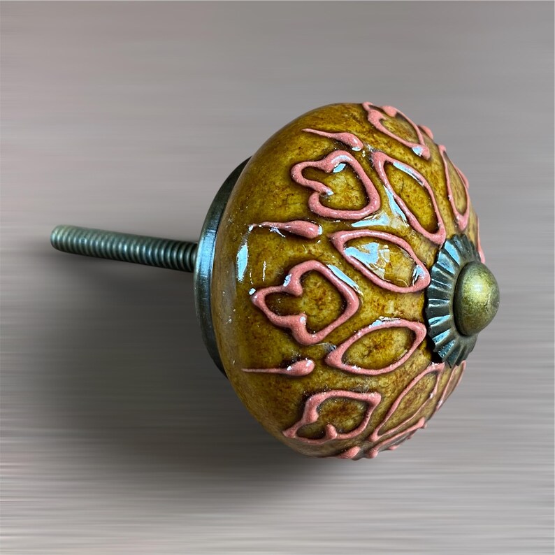 May include: A decorative cabinet knob with a mustard yellow base and pink floral design. The knob has a metal screw and a decorative metal base. The knob is approximately 3.8 cm in diameter.