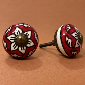 May include: Two round ceramic cabinet knobs featuring a red background with a white floral pattern and black outlines. Each knob has a central brass-toned metal cap and a threaded metal bolt extending from the back for installation purposes.