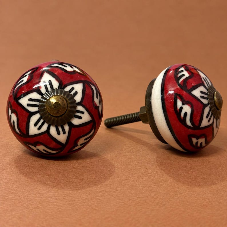 May include: Two round ceramic cabinet knobs featuring a red background with a white floral pattern and black outlines. Each knob has a central brass-toned metal cap and a threaded metal bolt extending from the back for installation purposes.