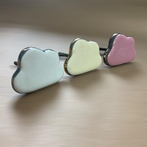 May include: Three cloud-shaped cabinet knobs in white, pale yellow, and pink. Each knob has a silver-coloured trim and a dark grey screw. The knobs are arranged on a light brown surface.