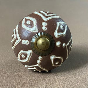 Puede incluir: Un pomo de armario de cer&aacute;mica marr&oacute;n con un patr&oacute;n geom&eacute;trico blanco. El pomo tiene un centro de color lat&oacute;n y un borde decorativo texturizado. Este herraje decorativo es adecuado para cajones y armarios.