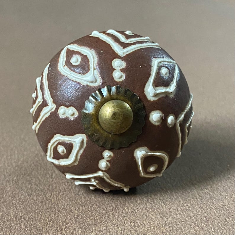 Puede incluir: Un pomo de armario de cer&aacute;mica marr&oacute;n con un patr&oacute;n geom&eacute;trico blanco. El pomo tiene un centro de color lat&oacute;n y un borde decorativo texturizado. Este herraje decorativo es adecuado para cajones y armarios.