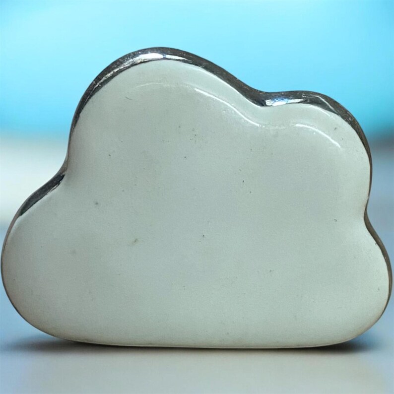 May include: A white cloud-shaped ornament with a silver-coloured trim. The cloud has a smooth, glossy surface and a rounded shape. The background is a light blue colour.