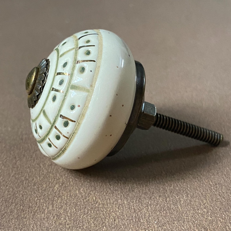Puede incluir: Pomo de armario de cer&aacute;mica color crema con un dise&ntilde;o decorativo en verde y dorado. El pomo tiene una base de metal y un perno roscado para la instalaci&oacute;n. El dise&ntilde;o presenta un patr&oacute;n repetitivo de l&iacute;neas y puntos.