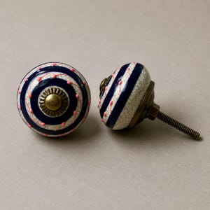 Op de afbeelding: Twee decoratieve keramische kastknoppen. Elke knop heeft een witte basis met marineblauwe strepen en roze stippen. De knoppen hebben een messingkleurige metalen basis en een schroef.