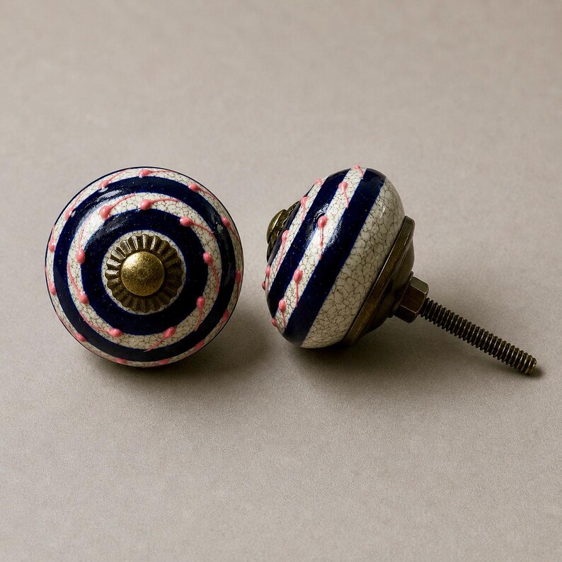 Op de afbeelding: Twee decoratieve keramische kastknoppen. Elke knop heeft een witte basis met marineblauwe strepen en roze stippen. De knoppen hebben een messingkleurige metalen basis en een schroef.