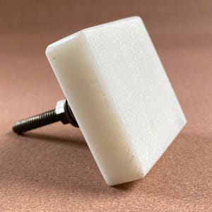 Pode incluir: Um puxador de arm&aacute;rio quadrado, branco sujo, com um parafuso de metal roscado. O puxador &eacute; feito de um material s&oacute;lido e opaco e tem uma superf&iacute;cie ligeiramente texturizada. O parafuso &eacute; cinza escuro e tem uma porca na base do puxador.