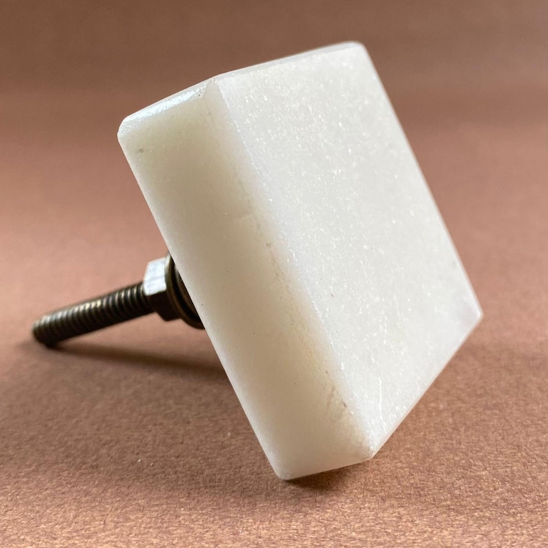 Pode incluir: Um puxador de arm&aacute;rio quadrado, branco sujo, com um parafuso de metal roscado. O puxador &eacute; feito de um material s&oacute;lido e opaco e tem uma superf&iacute;cie ligeiramente texturizada. O parafuso &eacute; cinza escuro e tem uma porca na base do puxador.