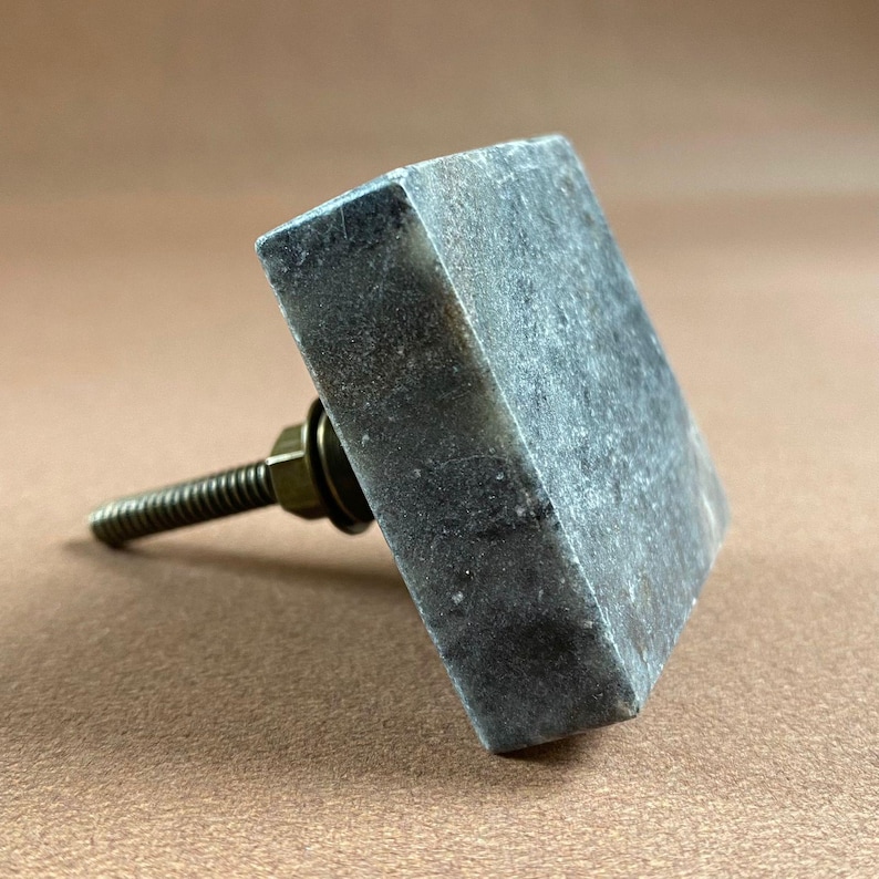 Pode incluir: Um puxador de arm&aacute;rio quadrado de m&aacute;rmore cinzento com um parafuso e uma arruela de cor lat&atilde;o. O puxador &eacute; feito de uma pedra s&oacute;lida e texturizada e foi concebido para ser usado em gavetas ou arm&aacute;rios. O hardware &eacute; de cor neutra.