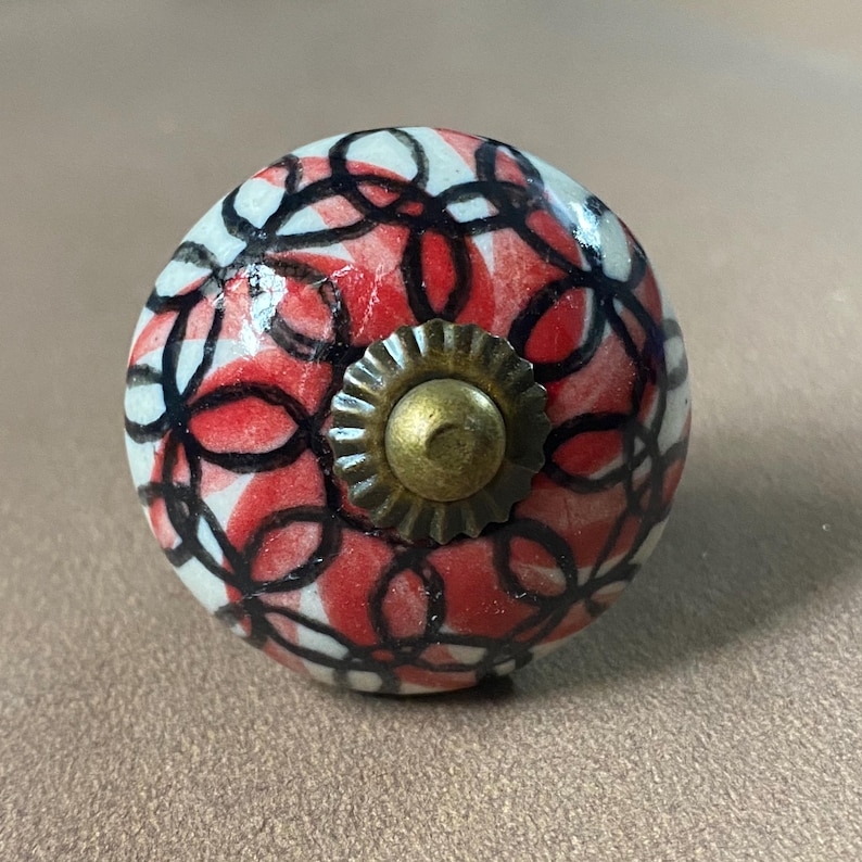 Puede incluir: Un tirador decorativo para armario con base blanca, con un patr&oacute;n de c&iacute;rculos rojos y negros superpuestos. El tirador tiene una base en forma de sol de color lat&oacute;n y un tornillo central. Este herraje &uacute;nico a&ntilde;ade un toque de color y estilo a cualquier mueble.