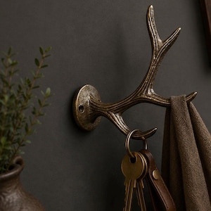 Könnte beinhalten: Bronze-farbener Wandhaken in Geweihform. Der Haken ist an einer dunklen Wand befestigt und hält ein braunes Handtuch und einen Schlüsselbund an einem Leder-Schlüsselanhänger. Der Haken hat eine rustikale, Vintage-Ästhetik.