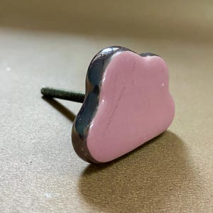 May include: A decorative cabinet knob with a pink, cloud-shaped ceramic face and a dark metallic border. The knob is attached to a threaded metal screw, suitable for furniture hardware. The knob is approximately 3.8 cm wide.