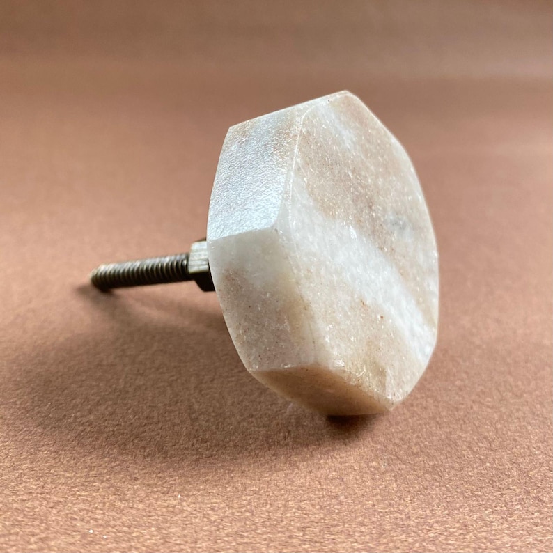 Pode incluir: Um puxador de arm&aacute;rio hexagonal de m&aacute;rmore de cor clara com um parafuso de metal roscado. O puxador tem um padr&atilde;o natural, com veios e um acabamento polido. O hardware &eacute; projetado para m&oacute;veis e decora&ccedil;&atilde;o de casa.