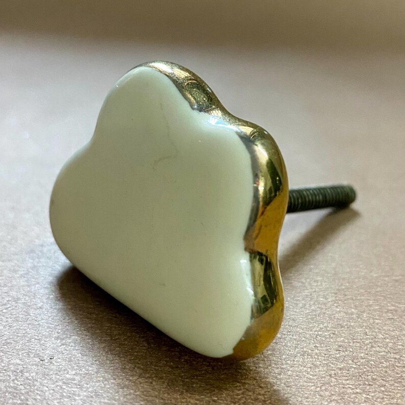 May include: A heart-shaped cabinet knob with a creamy white centre and gold-coloured trim. The knob has a threaded screw for easy installation. This decorative hardware adds a touch of elegance to furniture.