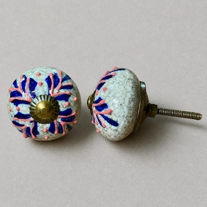 Op de afbeelding: Twee decoratieve keramische kastknoppen. De knoppen zijn wit met een gespikkelde textuur en hebben een blauw en roze bloemmotief. Elke knop heeft een messingkleurige basis en een schroefdraad voor installatie.