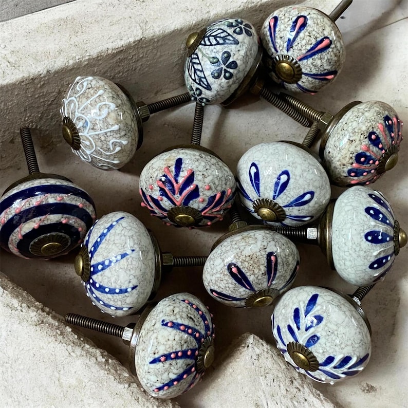 Op de afbeelding: Een verzameling decoratieve keramische kastknoppen. De knoppen hebben verschillende ontwerpen, waaronder bloempatronen, strepen en abstracte vormen, in kleuren als blauw, roze en wit. Elke knop heeft een metalen schroef voor bevestiging.