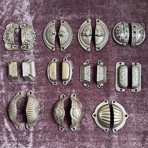 May include: A collection of twelve pairs of metal drawer pulls in various shapes and finishes, including antique brass, bronze, and silver tones. The hardware features diverse designs such as shell patterns, geometric engravings, and filigree details.