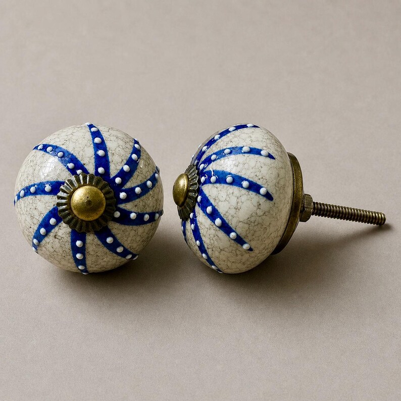 Op de afbeelding: Twee decoratieve keramische kastknoppen. De knoppen zijn gebroken wit met een gespikkeld patroon en versierd met blauwe strepen en witte stippen. Elke knop heeft een messingkleurige basis en een schroefdraad voor installatie.