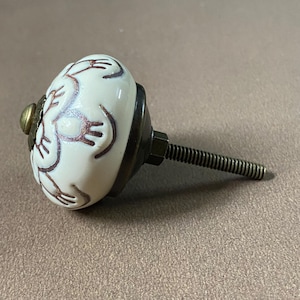 Puede incluir: Pomo de armario de cer&aacute;mica blanca con dise&ntilde;o floral marr&oacute;n. El pomo tiene una base color lat&oacute;n y un tornillo met&aacute;lico roscado para la instalaci&oacute;n. Herraje para cajones, armarios o muebles.