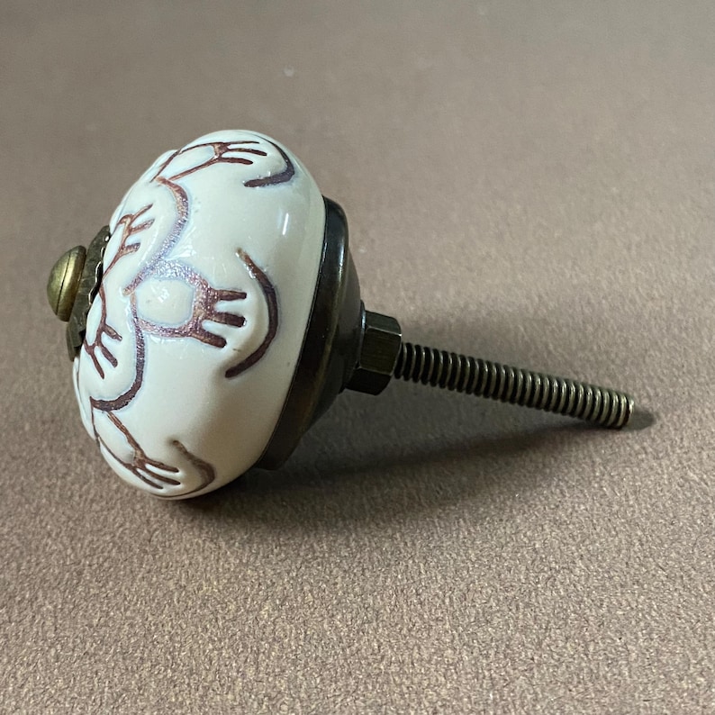Puede incluir: Pomo de armario de cer&aacute;mica blanca con dise&ntilde;o floral marr&oacute;n. El pomo tiene una base color lat&oacute;n y un tornillo met&aacute;lico roscado para la instalaci&oacute;n. Herraje para cajones, armarios o muebles.
