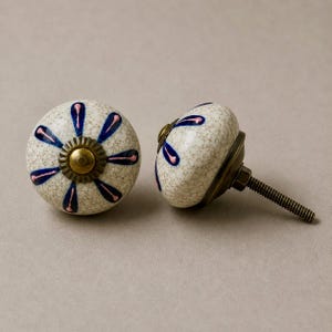 Op de afbeelding: Twee decoratieve keramische kastknoppen. De knoppen zijn gebroken wit met een craquel&eacute;-afwerking en hebben een bloemmotief in blauw en roze. Elke knop heeft een messingkleurige basis en een schroefdraadbout voor installatie.
